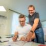 Ausbildungssituation im Büro: Eine Fachkraft und eine lernende Person arbeiten gemeinsam an technischen Zeichnungen und Projektunterlagen. Auf dem Schreibtisch liegen Pläne, Arbeitsmaterialien und digitale CAD‑Darstellungen, die eine strukturierte Pro