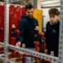 Ausbildungssituation in der Haustechnik: Eine erfahrene Fachkraft erklärt einer lernenden Person ein montiertes Rohrsystem mit Ventilen und Verbindungsstücken. Beide stehen in einer Werkstattumgebung mit Metallgestellen, Werkzeugen und technischen Anlag