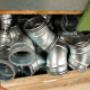 Das Bild zeigt eine Holzkiste voller metallischer Rohrbögen, die ordentlich übereinandergestapelt sind. Die silberfarbenen Formstücke bestehen aus verzinktem Metall und besitzen jeweils eine leichte Krümmung, wie sie typischerweise für Lüftungs‑ o