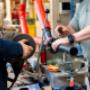 Auf dem Bild sind zwei Monteure zu sehen, deren Hände an einem professionellen Rohrschweißgerät arbeiten. Ein schwarzes Kunststoffrohr wird eingespannt und bearbeitet. Die Umgebung ist eine Werkstatt mit Werkzeugen, Bauteilen und Maschinen. Das Bild ze