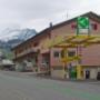 AGROLA Tankstelle in Amden. Gelbe Glasüberdachung mit einer Zanksäule und Zahlterminal. Digitaler Preisanzeige. Dahinter rotes Gebäude mit Volg und Bergkette im Hintergrund der Aufnahme.