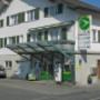 AGROLA Tankstelle in Einsiedeln. Mit kleiner Glasüberdachung. Zwei Tanksäulen und ein Zahlterminal. An Wohnhaus gelegen. Totem an Wohnhaus mit Preisanzeige und AGROLA Logo.