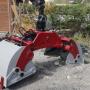 Ein robuster hydraulischer Greifer für Baumaschinen, farblich in Rot und Grau lackiert. Er steht auf einem Baustellengelände vor einer Holzhütte und ist bereit zum Einsatz.